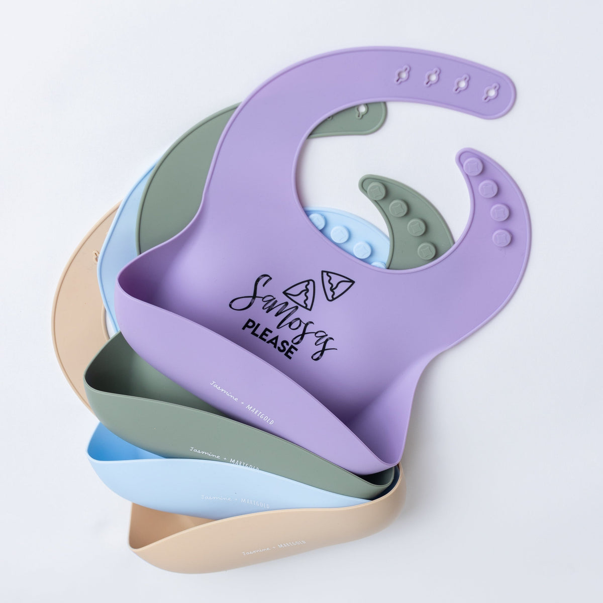 image of four bibs laying on top of each other.  Top color is lavendar, then olive green, then blue, then cream.  The bibs say "Samosas Please" with little samosas drawing.  They also have a white Jasmine + Marigold logo on them.image of four bibs laying on top of each other.  Top color is lavendar, then olive green, then blue, then cream.  The bibs say "Samosas Please" with little samosas drawing.  They also have a white Jasmine + Marigold logo on them.