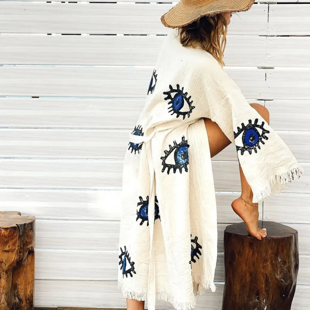The image shows a white kaftan-style robe made from 100% organic Turkish cotton. The robe features a relaxed fit with wide sleeves and a belted waist. It is adorned with embroidered eye motifs in vibrant colors, adding an artistic and unique touch. The fabric has a textured, slightly crinkled appearance, enhancing its casual and comfortable feel. Made in Turkey, this robe combines traditional craftsmanship with modern design elements.