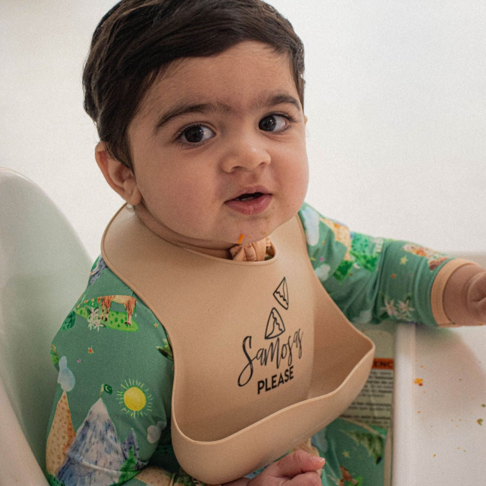 baby boy sitting in a high chair wearing a green onesie and a cream colored bib that says samosas please on it.  Baby has crumbs on his face.
