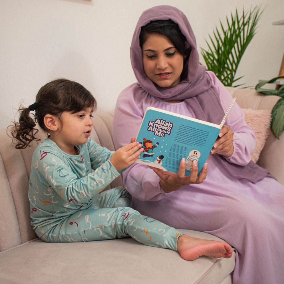 mother in purple dress and hijab reading a book to child on sofa with arabic pjs.