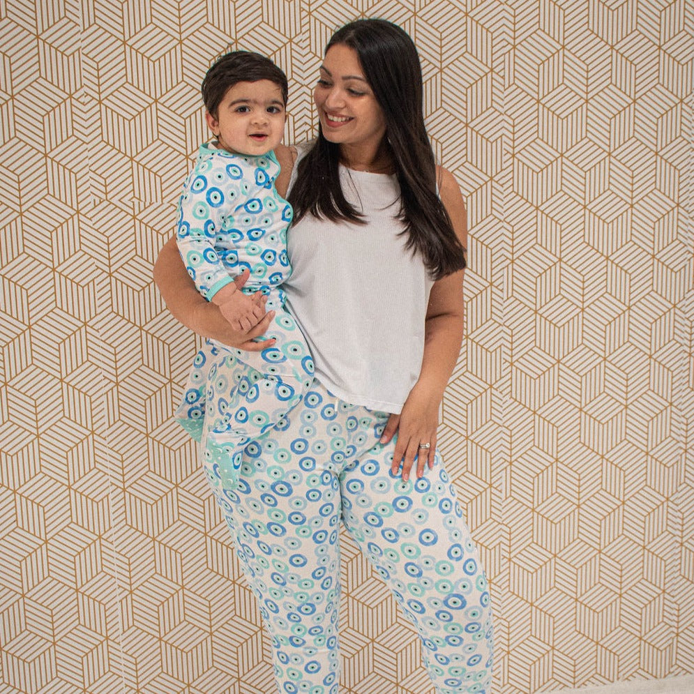 mom holding child on her hip, wearing blue joggers and a white tank top.  both are wearing evil eye print clothing.  background is patterned wallpapermom holding child on her hip, wearing blue joggers and a white tank top.  both are wearing evil eye print clothing.  background is patterned wallpaper