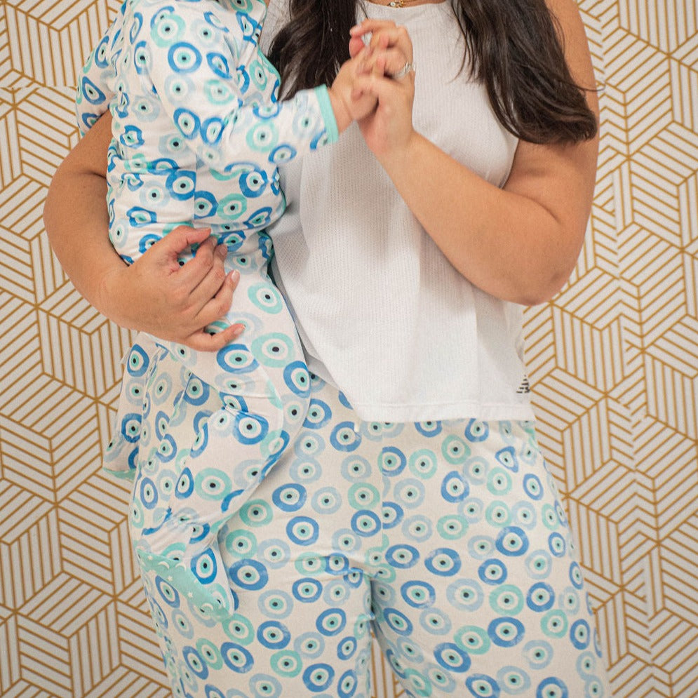 mom holding child on her hip, wearing blue joggers and a white tank top.  both are wearing evil eye print clothing.  background is patterned wallpaper