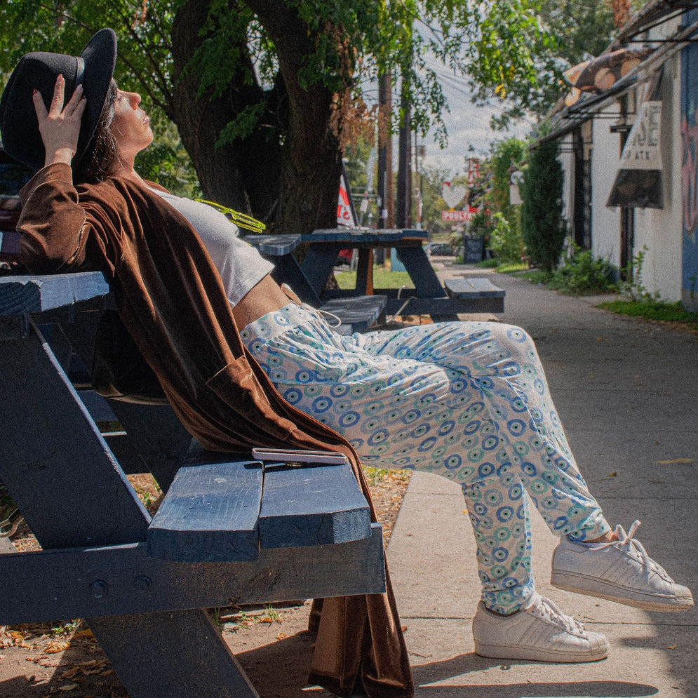 woman lounging on a bench outside with a long brown coat, black cap, and blue evil eye joggers.