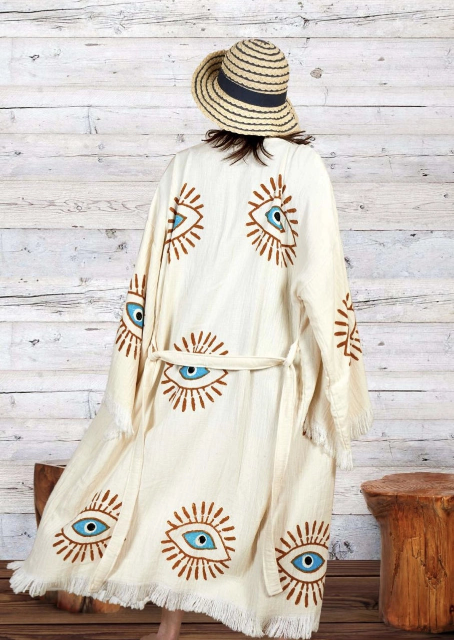The image shows a white kaftan-style robe made from 100% organic Turkish cotton. The robe features a relaxed fit with wide sleeves and a belted waist. It is adorned with embroidered eye motifs in vibrant colors, adding an artistic and unique touch. The fabric has a textured, slightly crinkled appearance, enhancing its casual and comfortable feel. Made in Turkey, this robe combines traditional craftsmanship with modern design elements.