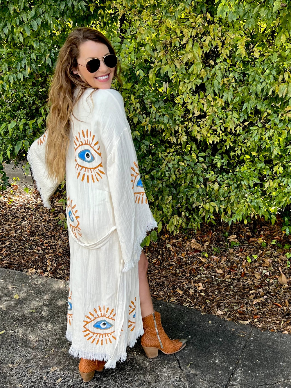The image shows a white kaftan-style robe made from 100% organic Turkish cotton. The robe features a relaxed fit with wide sleeves and a belted waist. It is adorned with embroidered eye motifs in vibrant colors, adding an artistic and unique touch. The fabric has a textured, slightly crinkled appearance, enhancing its casual and comfortable feel. Made in Turkey, this robe combines traditional craftsmanship with modern design elements.