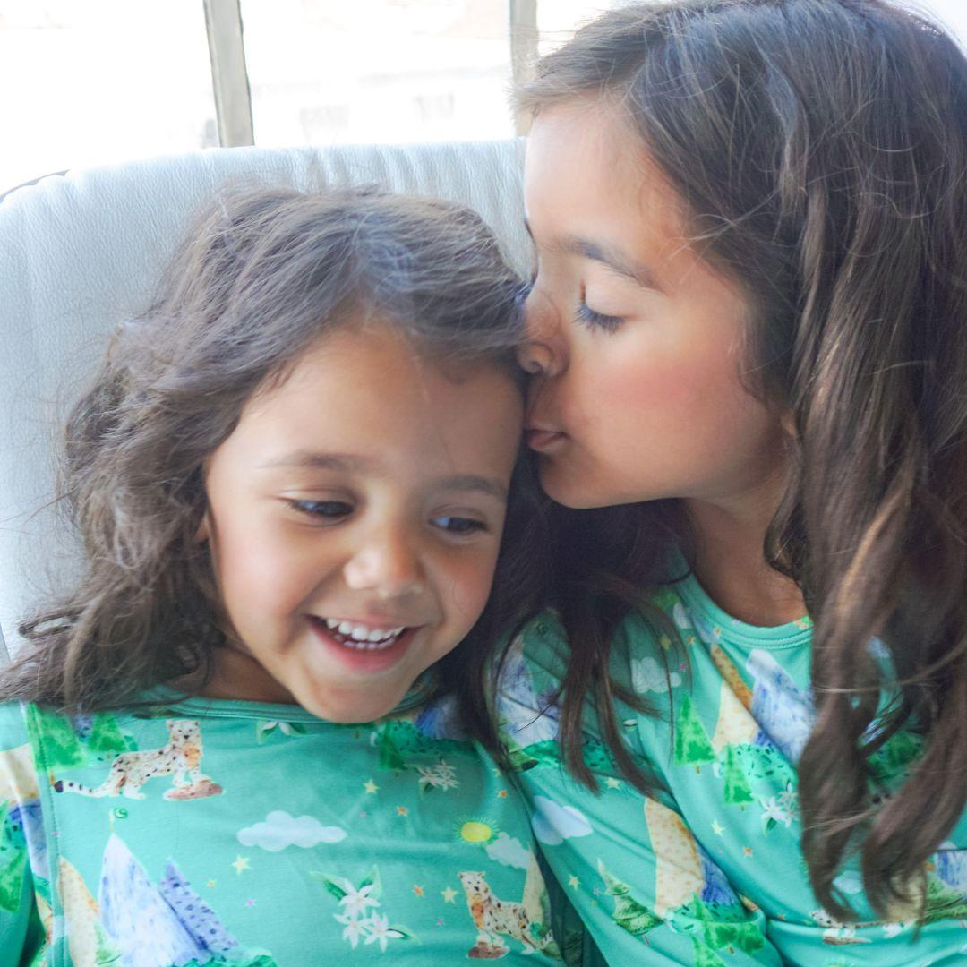 two girls sitting on a sofa laughing and giggling.  older girl kissing younger girl on forehead.  pajama print is green background.