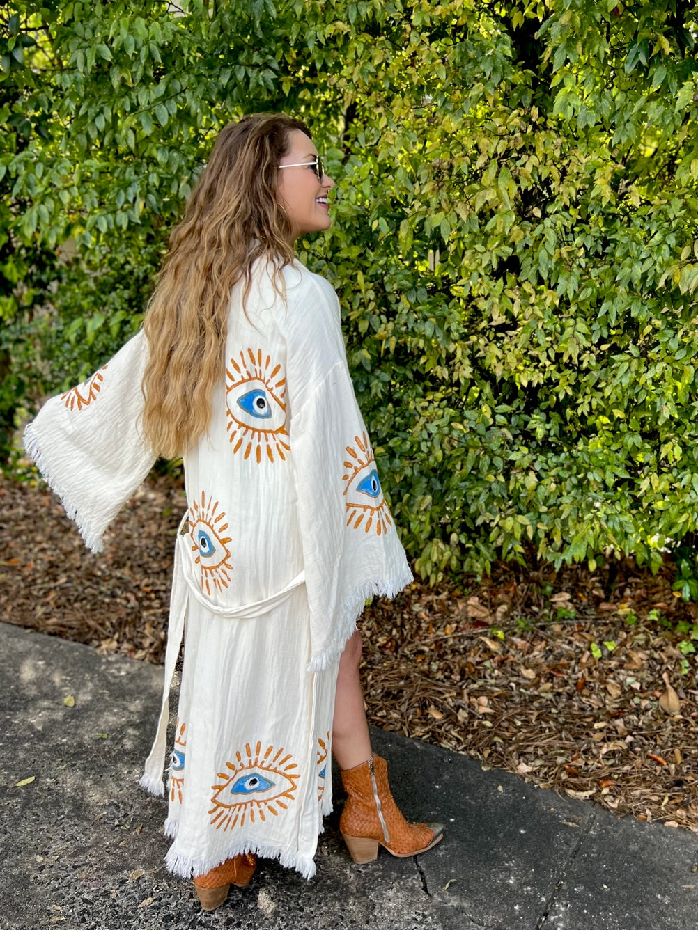 The image shows a white kaftan-style robe made from 100% organic Turkish cotton. The robe features a relaxed fit with wide sleeves and a belted waist. It is adorned with embroidered eye motifs in vibrant colors, adding an artistic and unique touch. The fabric has a textured, slightly crinkled appearance, enhancing its casual and comfortable feel. Made in Turkey, this robe combines traditional craftsmanship with modern design elements.