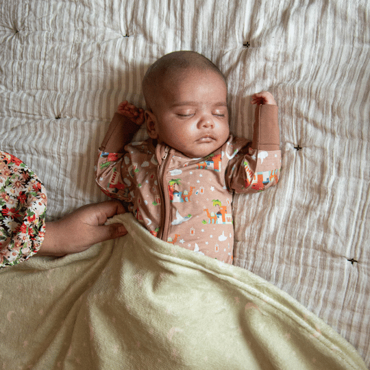 A baby sleeping soundly in a Sleeper Pajama with a pattern that includes a depiction of a desert town, arched windows, and date palm trees.