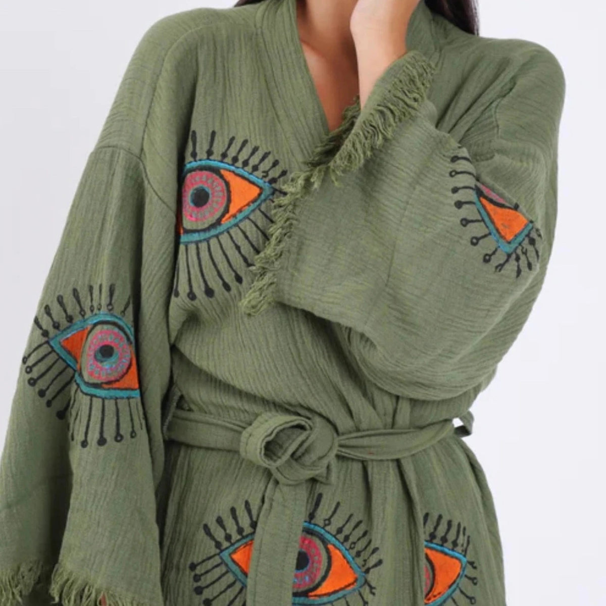 The image shows a green kaftan-style robe made from 100% organic Turkish cotton. The robe features a relaxed fit with wide sleeves and a belted waist. It is adorned with embroidered eye motifs in vibrant colors, adding an artistic and unique touch. The fabric has a textured, slightly crinkled appearance, enhancing its casual and comfortable feel. Made in Turkey, this robe combines traditional craftsmanship with modern design elements.