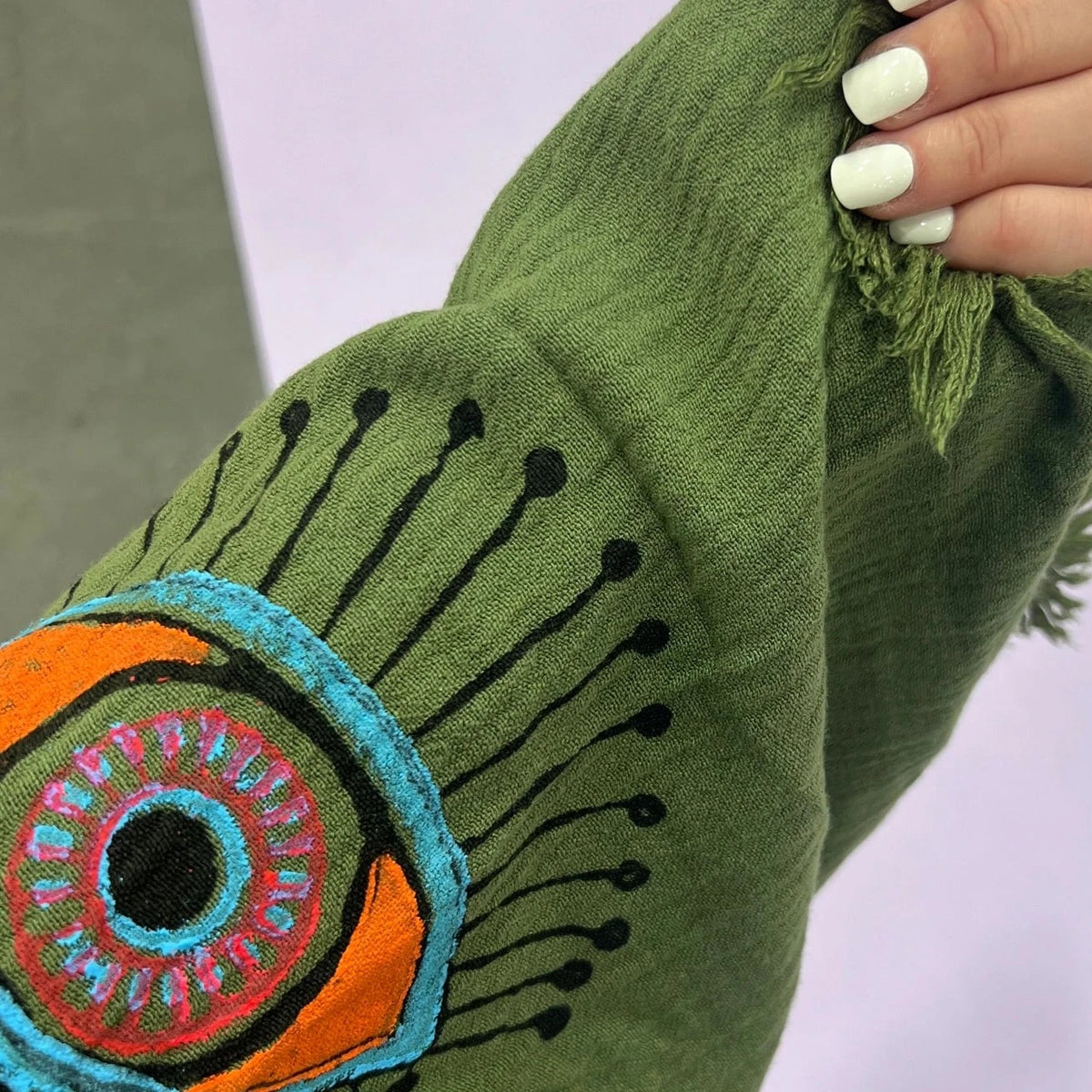 The image shows a green kaftan-style robe made from 100% organic Turkish cotton. The robe features a relaxed fit with wide sleeves and a belted waist. It is adorned with embroidered eye motifs in vibrant colors, adding an artistic and unique touch. The fabric has a textured, slightly crinkled appearance, enhancing its casual and comfortable feel. Made in Turkey, this robe combines traditional craftsmanship with modern design elements.